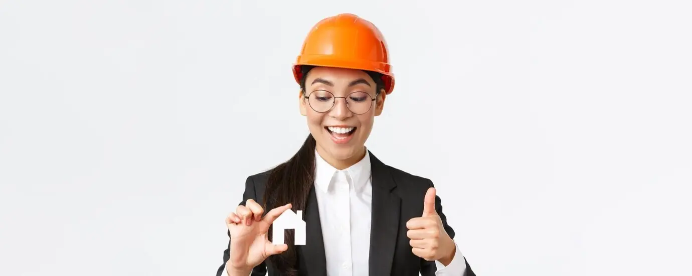 Close-up of a happy, pleased Asian female architect/construction engineer wearing a helmet and suit, looking at a miniature house and giving a thumbs-up to guarantee the quality of repair works, on a white background.