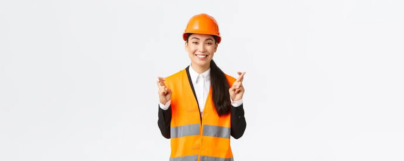 Smiling, hopeful, and optimistic Asian female engineer and construction manager wearing a safety helmet, crossing her fingers for good luck and showing faith in the project, looking positive while standing against a white background.